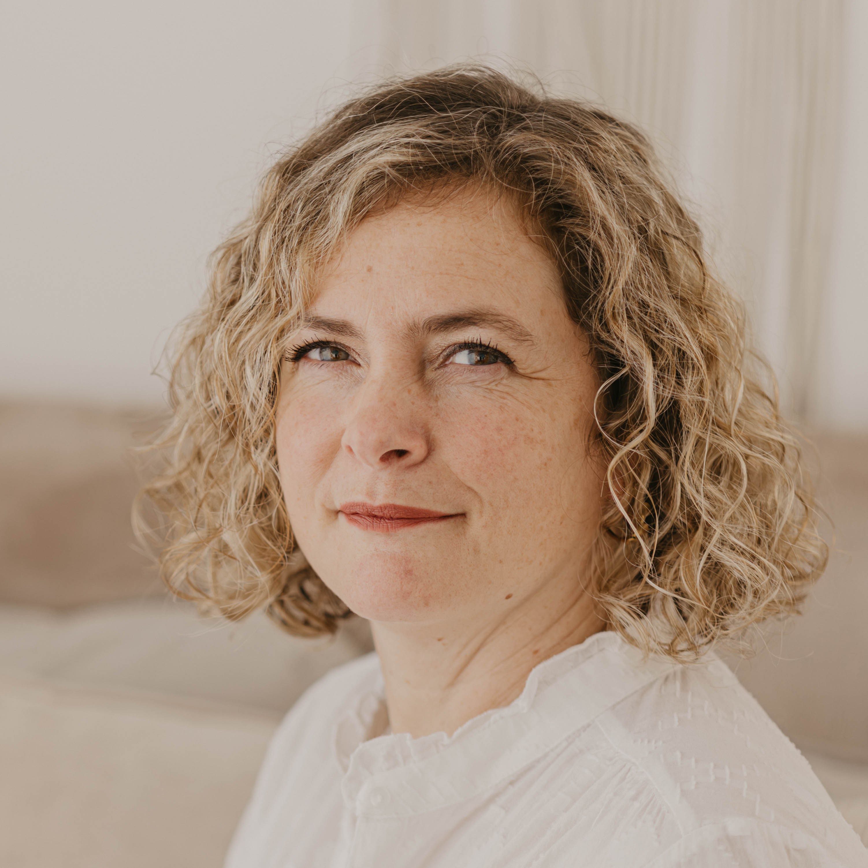 Woman with curly blonde hair and a white blouse smiling softly, sitting in a neutral-toned room.